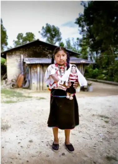 Niña con vestimenta tradicional sosteniendo una muñeca artesanal frente a una casa de campo.