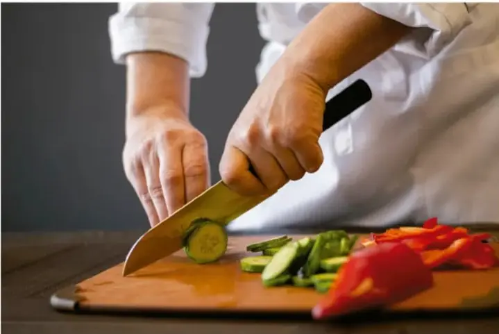 Fotografía de una persona (manos y torso con delantal/chamarra blanca) cortando rodajas de pepino y tiras de pimiento rojo sobre una tabla de cocina de madera con un cuchillo.