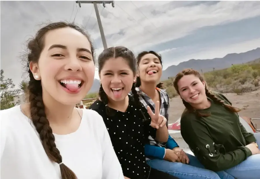 Fotografía de cuatro jóvenes sonriendo en un recorrido en camioneta; muestran distintos tipos de cabello: una trenza larga, cabello rizado corto con coletas, cabello ondulado y trenzas, con montañas y cielo nublado al fondo.