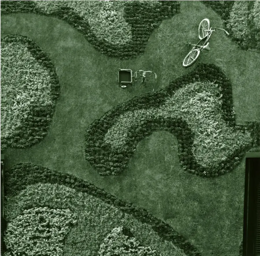 Fotografía aérea de un jardín: sobre el césped se dibujan grandes motivos orgánicos formados por matas de plantas de distintas alturas y tonos; en los senderos de pasto aparecen una bicicleta y un triciclo de carga. Crédito al pie: Alejandra García Torres, grupo de México.
