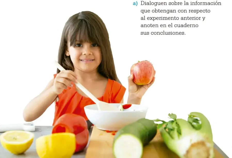 Niña sosteniendo una manzana y una cuchara, con frutas y verduras sobre la mesa.