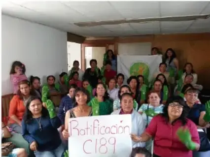 Grupo de personas, en su mayoría mujeres, sostienen un cartel que dice 'Ratificación C189', en un evento o reunión.