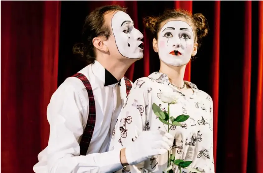 Fotografía de dos actores con maquillaje blanco tipo payaso/mimo en escenario, con cortinas rojas de fondo; uno susurra al oído de la otra, quien sostiene una rosa blanca. Ilustra la representación teatral en vivo.