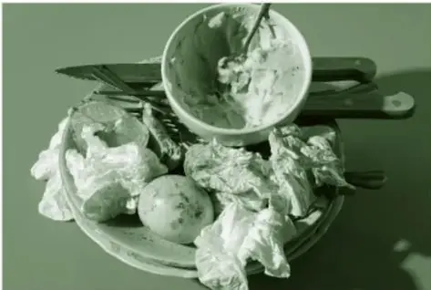 Fotografía en tono verde de un bote de basura desbordado con residuos: restos de comida, envolturas, un plato y utensilios amontonados.