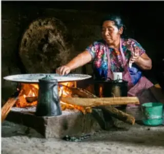 Mujer indígena moliendo maíz en un metate junto a un fogón tradicional.