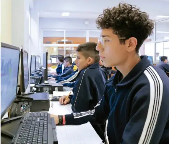 Fotografía de estudiantes de telesecundaria trabajando en computadoras dentro de un aula o laboratorio de cómputo, sentados frente a monitores y teclados.