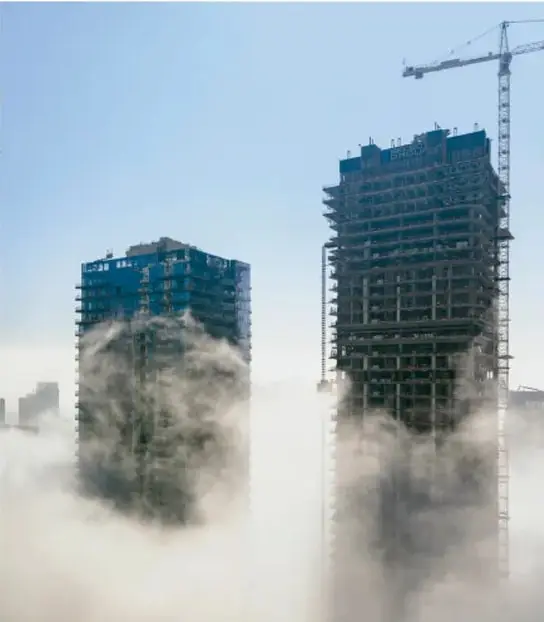 Fotografía de dos edificios en construcción rodeados de niebla o esmog, con una grúa de fondo, que ilustra el impacto de la actividad humana en el ambiente.