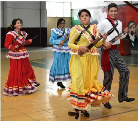 Jóvenes estudiantes con trajes regionales de colores (rojo, azul, amarillo) sosteniendo armas de utilería, posando en lo que parece un gimnasio escolar, representando una escena de la Revolución Mexicana.