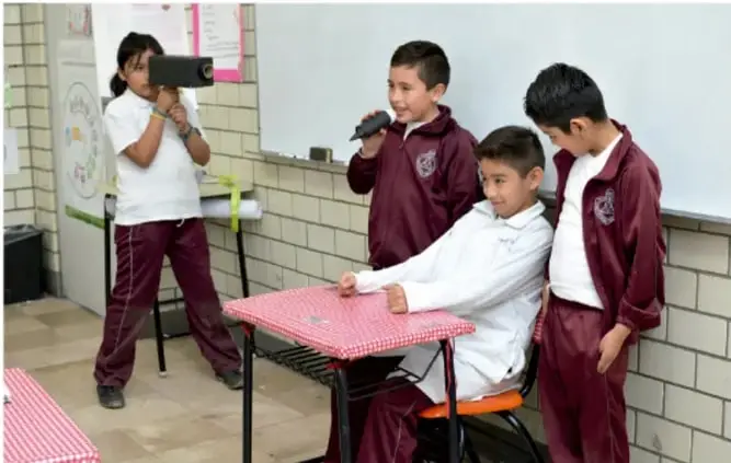Fotografía de un grupo de estudiantes de primaria en un salón de clases. Una niña sostiene una cámara de video y graba a un compañero sentado en una silla de ruedas frente a una mesa con mantel rojo; otros dos compañeros observan de pie. Representa la inclusión y el trabajo colaborativo del proyecto de revista escolar.