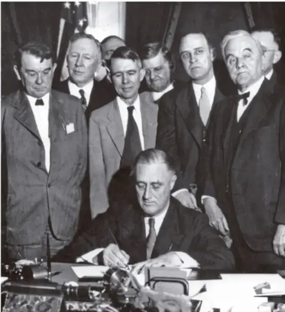 Fotografía en blanco y negro de Franklin D. Roosevelt firmando documentos rodeado de varios hombres con traje.
