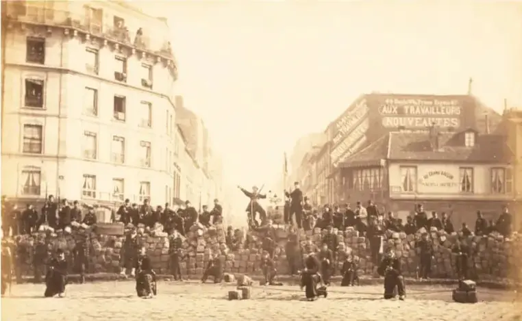 Fotografía histórica en sepia que muestra una calle de París durante la Comuna, con una multitud reunida, barricadas y edificios con letreros como 'AUX TRAVAILLEURS'.