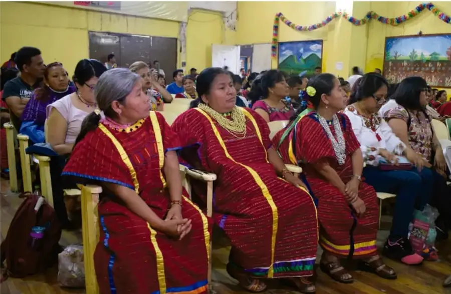 Fotografía de mujeres indígenas vestidas con huipiles rojos tradicionales sentadas en filas dentro de un salón, asistiendo a una asamblea o acto comunitario.
