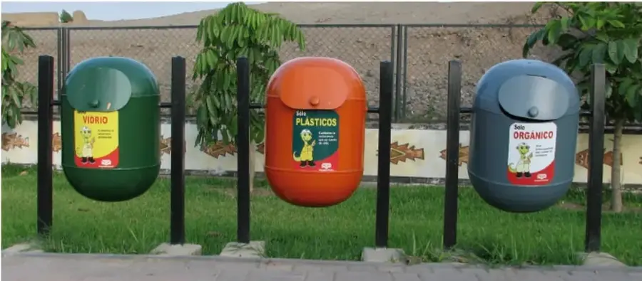 Tres contenedores de reciclaje colgados de postes: verde para vidrio, naranja para plásticos y gris para orgánico, ubicados en un parque con pasto y árboles.