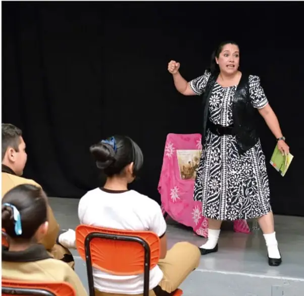 Fotografía de una mujer con vestido indígena ante un grupo de alumnos sentados, aparentemente en una presentación o clase teatral con fondo de telón negro.