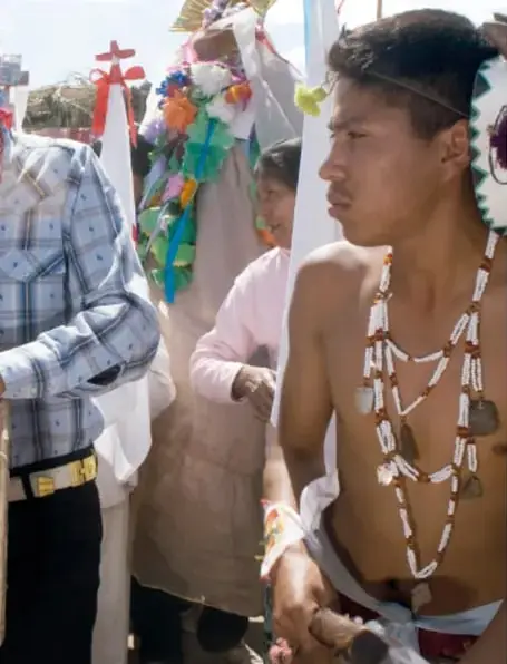 Músicos de la danza: un hombre con torso desnudo, collares blancos de cuentas y sonaja, y otro músico con camisa de cuadros tocando una flauta de carrizo, entre banderas y adornos de la fiesta.